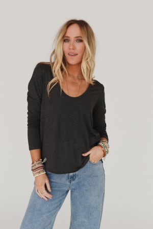 A woman with long blonde hair poses against a plain white background wearing the Ryder Long Sleeve Bralette Tee in Charcoal, light blue jeans, layered necklaces, and multiple bracelets.
