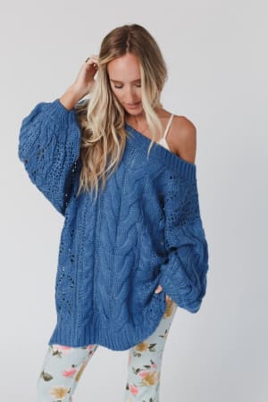 A woman with long blonde hair wears the Golden Hour Bubble Sleeve Sweater Dress in blue, standing against a plain white background. She looks down and touches her hair with one hand.