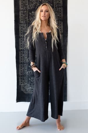 A woman with long blonde hair stands barefoot before a black tapestry, wearing the Simply Comfort Jumpsuit in dark gray—an oversized, long-sleeved boho style with wide legs, a button-up front, and pockets for a relaxed look.