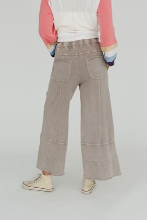 A person stands with their back to the camera against a plain background, wearing the Lucky One Wide Leg Pant in taupe, a long-sleeve colorblock top with pink, white, and navy sleeves, and white sneakers.