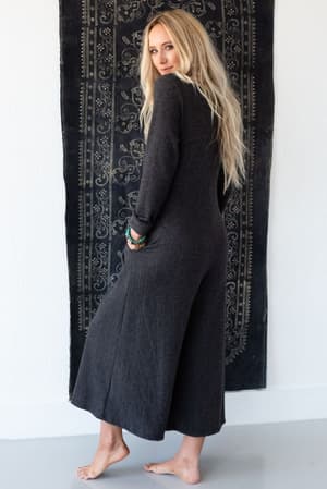 A barefoot woman with long blonde hair wears the Simply Comfort Jumpsuit in dark gray, featuring a loose, oversized boho silhouette. She stands before a patterned black tapestry, glancing over her shoulder at the camera.