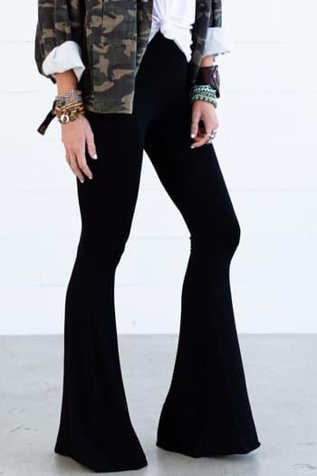 Wearing Cher Solid Raw Edge Flare Pant in black, a person stands against a white wall. The photo is cropped to display only the flared pants and lower body, styled with a tied white shirt, camouflage jacket, and bracelets.