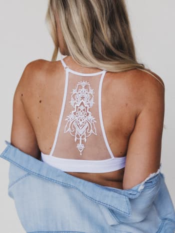 A woman with long blonde hair faces away, wearing the Tattoo Bralette - White featuring intricate lace on the back and a light blue denim jacket draped over her arms. The background is plain and neutral.