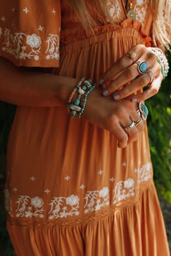 A person wearing an orange dress with white floral embroidery, turquoise rings, and the San Antonio Bracelet Set - Turquoise stands with hands folded, showcasing their boho bracelet style and manicured nails.