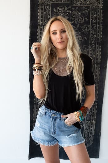 A woman with long blonde hair wears the Scoop Neck Bralette Tee in black, paired with ripped denim shorts and multiple bracelets. She stands before a decorative dark tapestry, showcasing effortless boho style.
