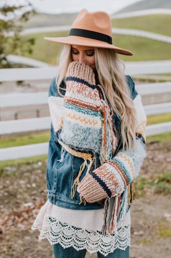 A woman with long blonde hair stands outdoors near a white fence and grassy hills, wearing the Sukie Sweater Denim Jacket - Blue and a tan hat, covering her mouth with her chunky knit sweater sleeve.