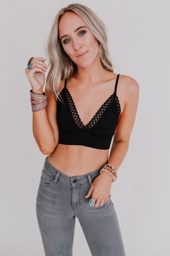 A woman with long blonde hair wears the Audra Waistband Loop Lace Bralette in black with adjustable straps and gray high-waisted jeans. She stands against a plain white background, confidently looking at the camera while wearing bracelets and rings.