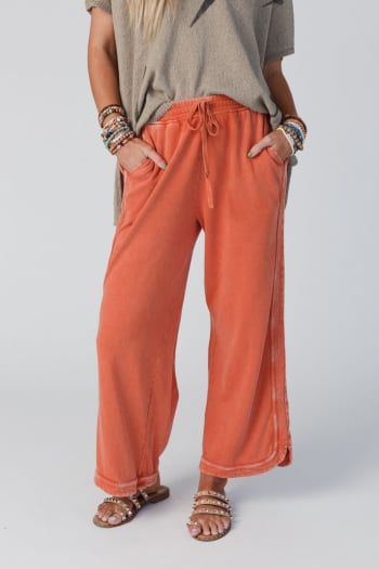 A person wearing So Comfy Wide Leg Pant Cropped Length - Faded Rust with a beige short-sleeve top, beaded bracelets, and tan studded sandals stands with hands in pockets. The face is not visible.