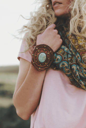 A woman with long curly blonde hair and a light pink t-shirt wears a colorful scarf, showing off the Aegean Boho Leather Cuff Bracelet - Brown on her wrist.