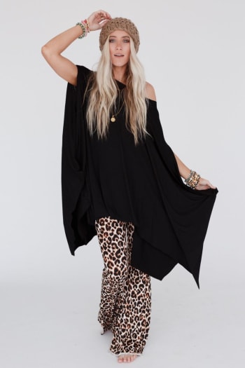 A woman with long blonde hair poses against a plain background wearing The Wren Tunic - Black, leopard print flared pants, a knitted beige hat, and multiple bracelets for a relaxed, stylish look.