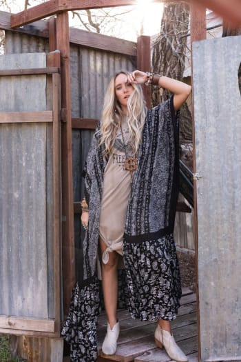 A woman with long blonde hair stands outdoors on wooden steps, wearing a beige dress and the Paisley Tapestry Free Flow Duster Kimono in black, styled with ankle boots and stacked bracelets as she poses with one hand on her forehead.
