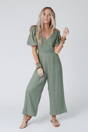 A woman with long, wavy blonde hair wears the Weekender Puff Sleeve Jumpsuit in olive, featuring wide legs. She accessorizes with layered necklaces, several bracelets, and beaded sandals while standing against a plain white background.