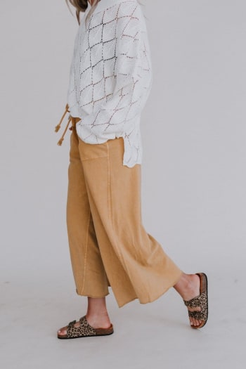 A person in a white patterned sweater, Relaxing Robin Wide Leg Pant - Mustard, and leopard print slide sandals walks against a plain light grey background. Their face is not visible.