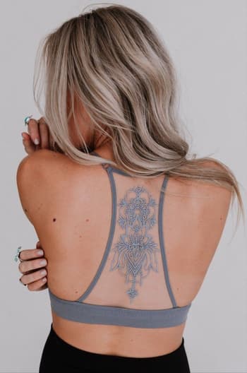 A woman with long, wavy blonde hair faces away, showing a mandala-style spine tattoo. She wears the Tattoo Bralette - Gray and rings on her fingers. The background is plain and light-colored.