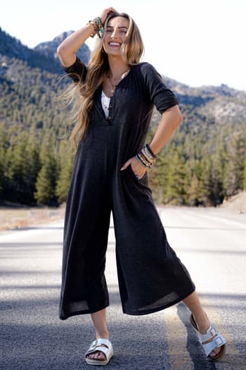 A smiling woman stands on a sunny mountain road in the Simply Comfort Jumpsuit - Dark Gray, a scoop neck boho style with an oversize silhouette. She pairs it with white sandals and colorful bracelets, framed by pine trees and rolling hills.