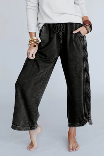 A person stands barefoot, hands in pockets, wearing a white long-sleeve top and the So Comfy Wide Leg Pant Cropped Length - Charcoal with a high-rise elastic waist, accessorized with multiple bracelets on both wrists.