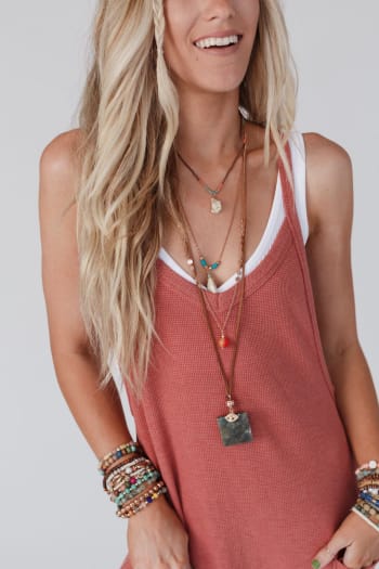 A smiling woman with long blonde hair wears a sleeveless rust-colored top over a white tank and the Giselle Stone and Shell Boho Necklace in Turquoise, paired with several colorful beaded bracelets on both wrists.
